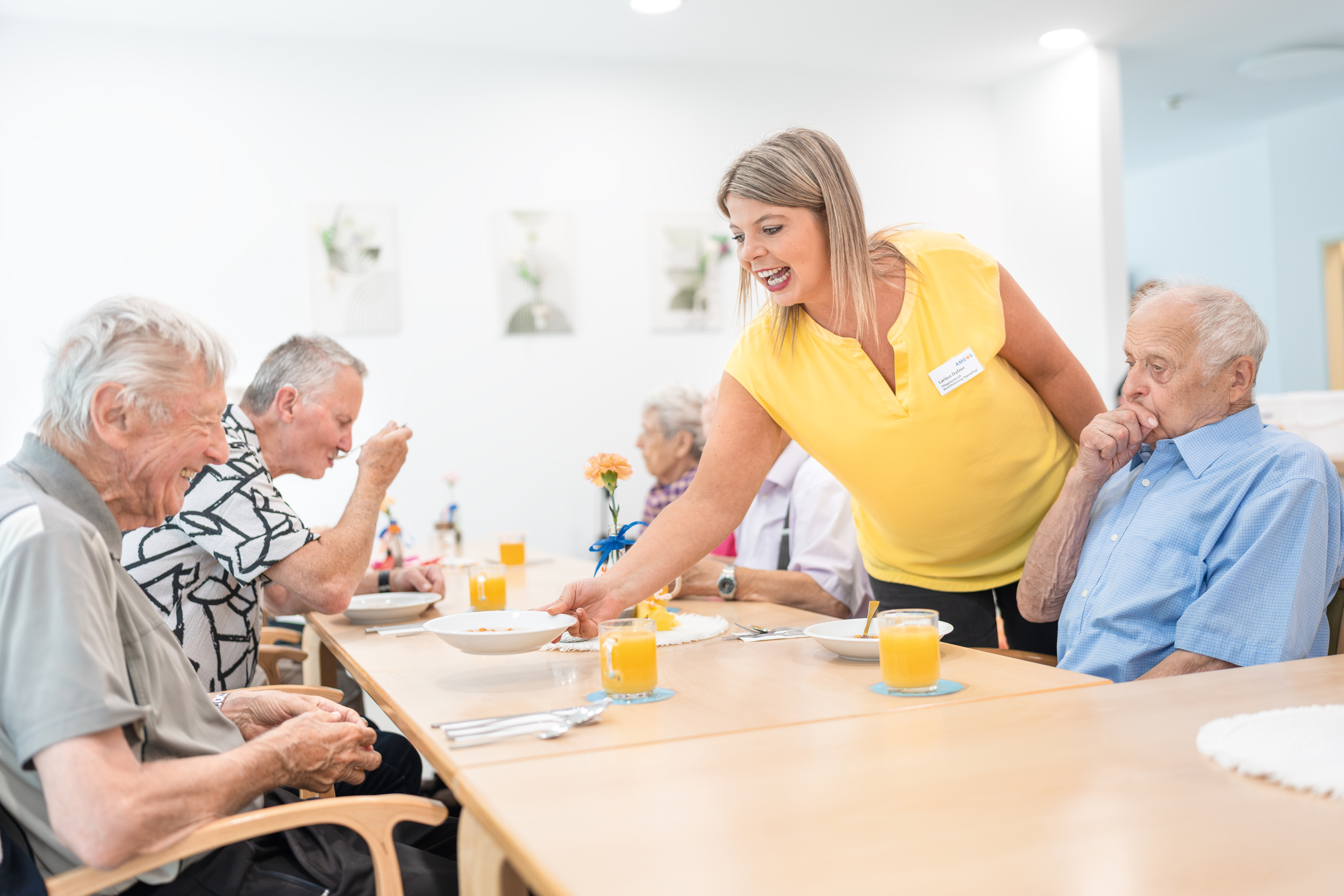 Gemeinsames Essen Drei ältere Herren sitzen am Esstisch, während eine Mitarbeiterin einen Teller mit Essen überreicht