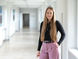 Frau mit dunklem Oberteil steht in einem Flur, eine Hand in der Tasche und lächelt in die Kamera.