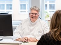 Arzt in Kittel sitzt am Schreibtisch vor einer Patientin und lächelt in die Kamera