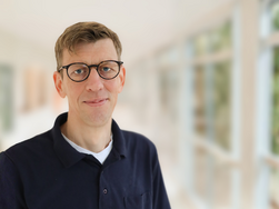 Dr. Claus Bolling, Chefarzt der Klinik für Onkologie, Gynäkologie und Urologie im AMEOS Reha Klinikum Ratzeburg blick lächelnd in die Kamera. Der Hintergrund ist schlicht, warm und hell (© AMEOS).