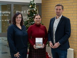 Zwei Frauen und ein Mann stehen vor einem Weihnachtsbaum und lächeln in die Kamera. Die Frau in der Mitte hält eine kleine Box mit Geld in der Hand.